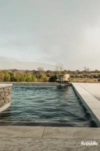 A serene swimming pool with a stone edge, featuring a modern water feature, surrounded by lush greenery and open landscapes
