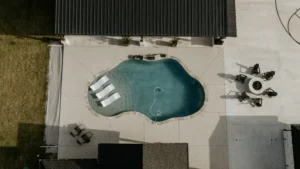 Aerial view of a backyard featuring a pool, four sun loungers, and a fire pit surrounded by black chairs on a patio