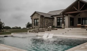 Modern stone house beside a swimming pool with submerged lounge chairs, set against a cloudy sky and lush green landscape