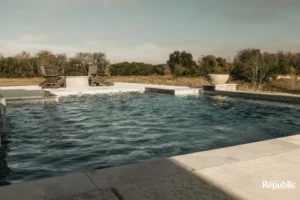 A serene swimming pool surrounded by two wooden chairs and a stone fountain, set against a tranquil outdoor landscape